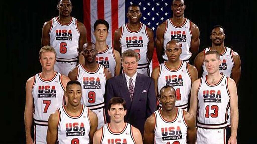 Dream Team Jogadores da seleção americana de basquete, com uniforme branco, ao redor de seu técnico e na frente da bandeira dos EUA.