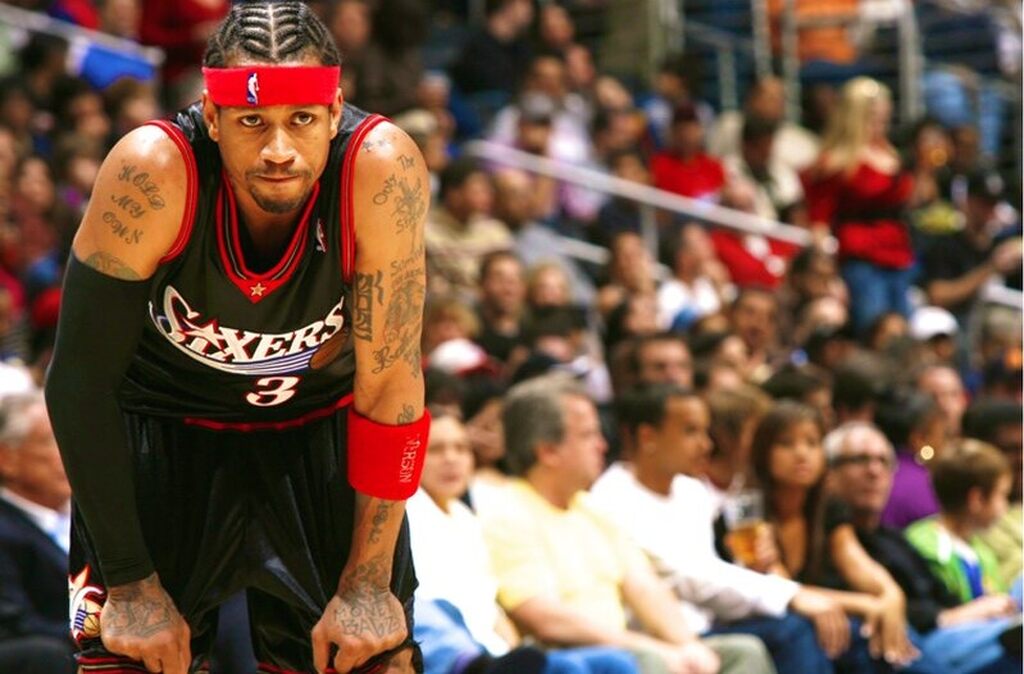 Ícones das quadras e da moda Jogador de basquete de uniforme preto com detalhes vermelhos, apoiado nos joelhos, cansado, durante jogo.