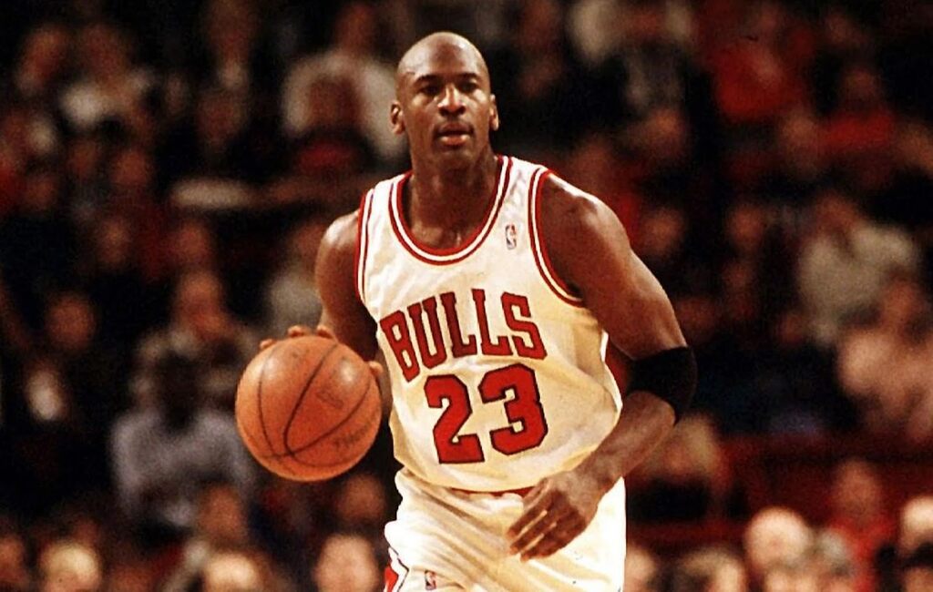 O maior de todos os tempos e os maiores do presente Jogador de basquete com uniforme branco com letras e detalhes em vermelhos, batendo bolando e progredindo pela quadra.