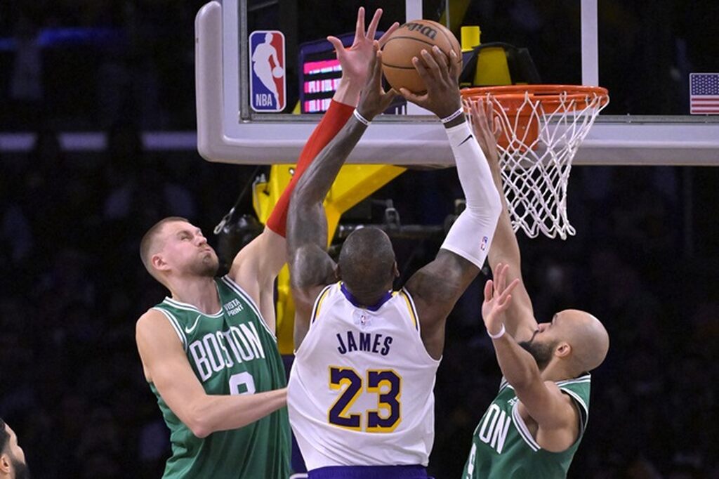 Jogador de basquete de uniforme branco com detalhes pretos tentando marcar um ponto, enquanto o adversário tenta bloquear.