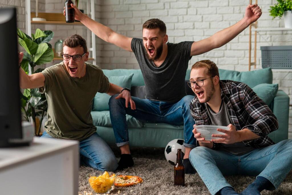 3 homens comemorando ago enquanto assistem TV, comem salgadinho e bebem cerveja, com uma bola perto.