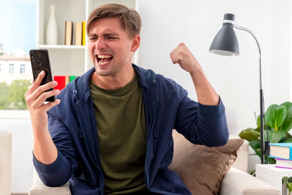 Homem de camisa verde e casaco azul, segurando um celular com a mão direita e comemorando algo.