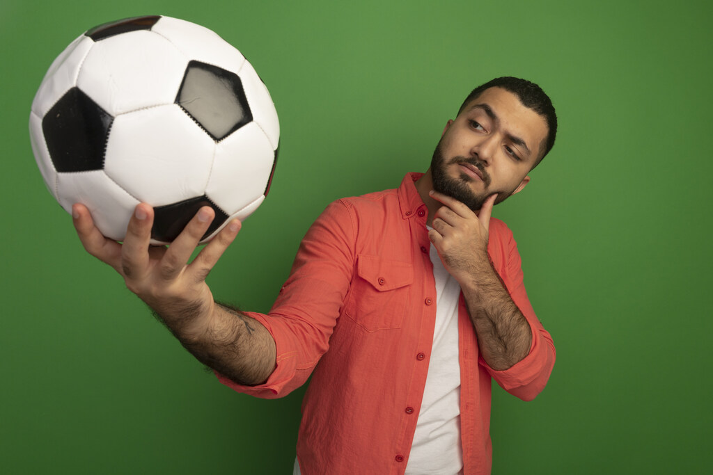 Homem de camisa branca e blusa rosa, em fundo verde, segurando uma bola de futebol e olhando pensativo pra ela.