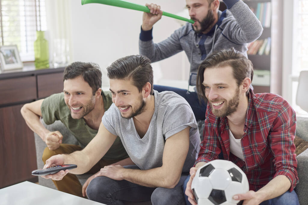 4 homens ao redor de sofá, assistindo ao jogo e torcendo intensamente, um deles com uma bola na mão.