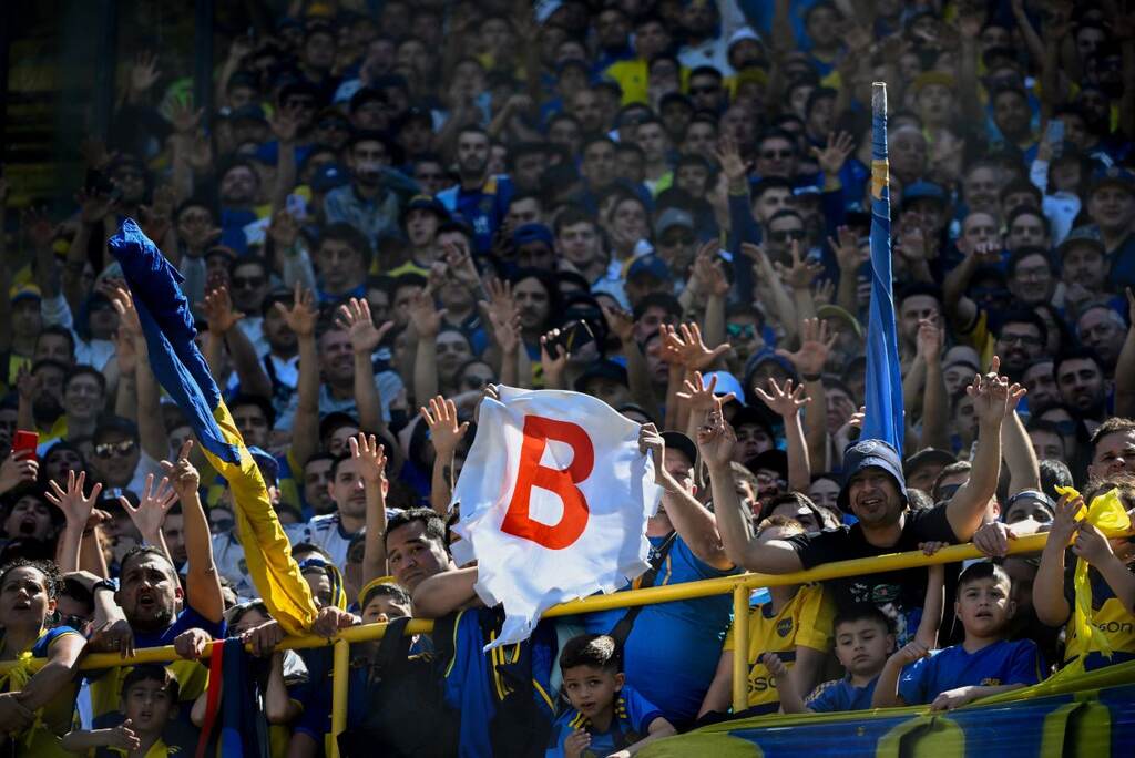 Torcedores com as cores azul e amarelo levantando uma bandeira com a letra "B" escrita em vermelho.