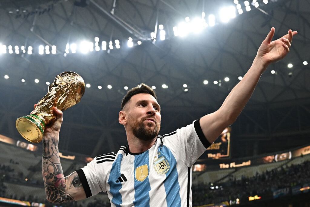 Jogador de futebol Lionel Messi, vestindo uniforme azul e branco da Argentina e erguendo taça da Copa do Mundo.