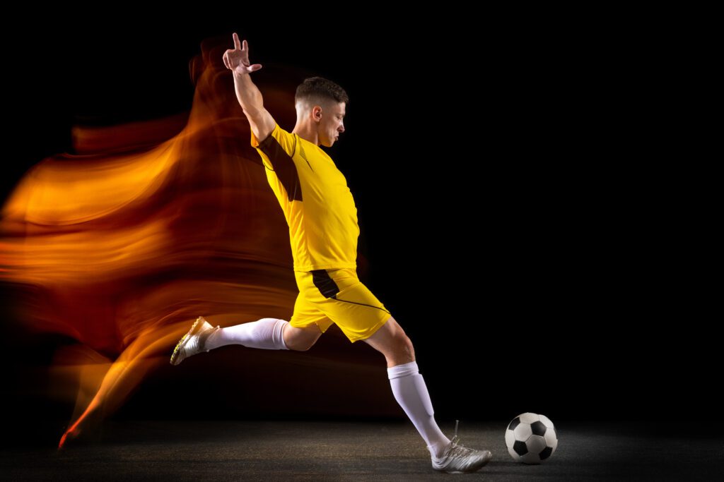 Jogador de futebol vestindo uniforme amarelo com detalhes pretos, chutando uma bola em um fundo escuro.