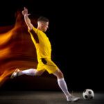 Jogador de futebol vestindo uniforme amarelo com detalhes pretos, chutando uma bola em um fundo escuro.