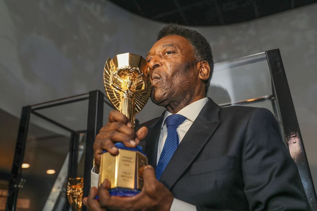 Rei do futebol, Pelé, segurando e beijando a antiga taça da Copa do Mundo, usando terno e gravata.