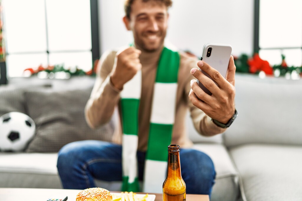 Homem de cachecol verde e branco, sentado em sofá om uma bola de futebol, segurando um celular e comemorado.