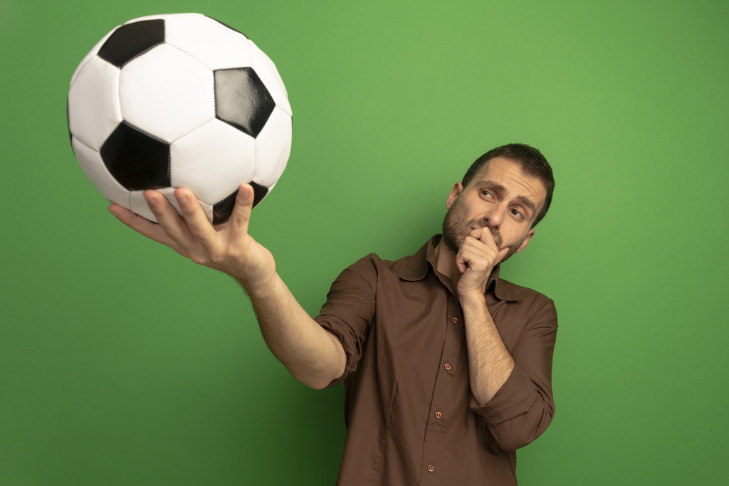 Homem de camisa marrom, pensativo, segurando uma bola de futebol e olhando para ela, em um fundo verde.