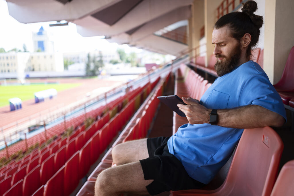 Homem de camisa azul e bermuda preta, pensativo e usando um tablet na arquibancada de um estádio de futebol vazio.