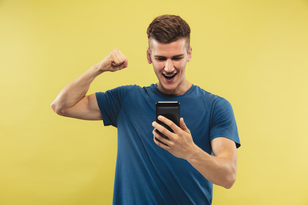 Homem de camisa azul, segurando um celular com a mão esquerda e comemorando em um fundo amarelo.