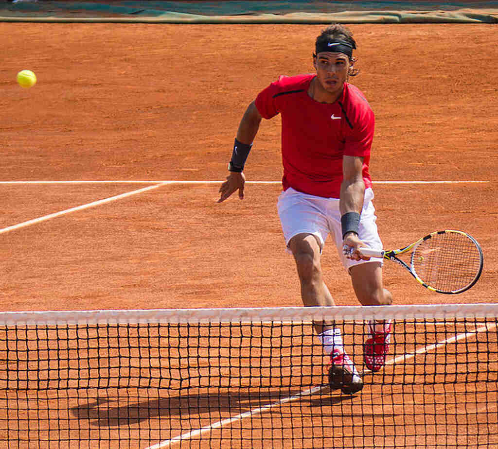 Técnica e estilo de jogo Jogador de tênis Rafael Nadal, vestindo uniforme vermelho e bermuda branca, correndo em direção a bola durante partida.