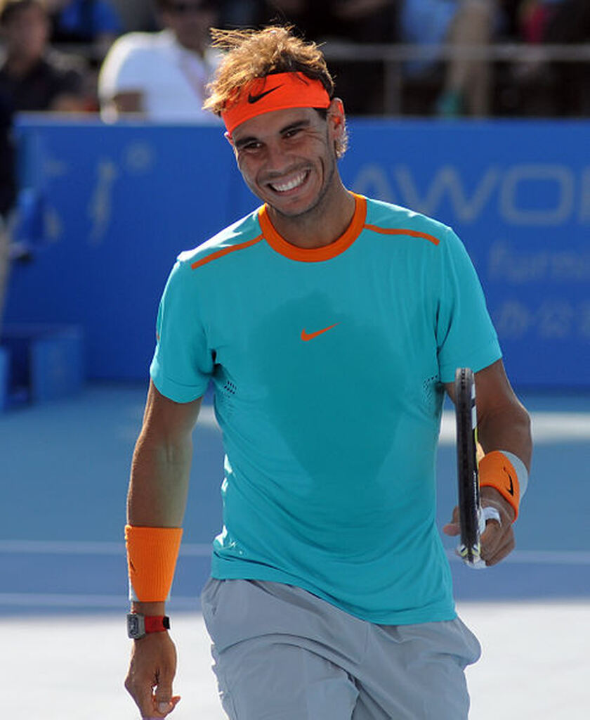 Ascensão no cenário do tênis mundial Jogador de tênis Rafael Ndal, vestindo uniforme azul e bermuda cinza, andando sorrindo pela quadra durante partida.