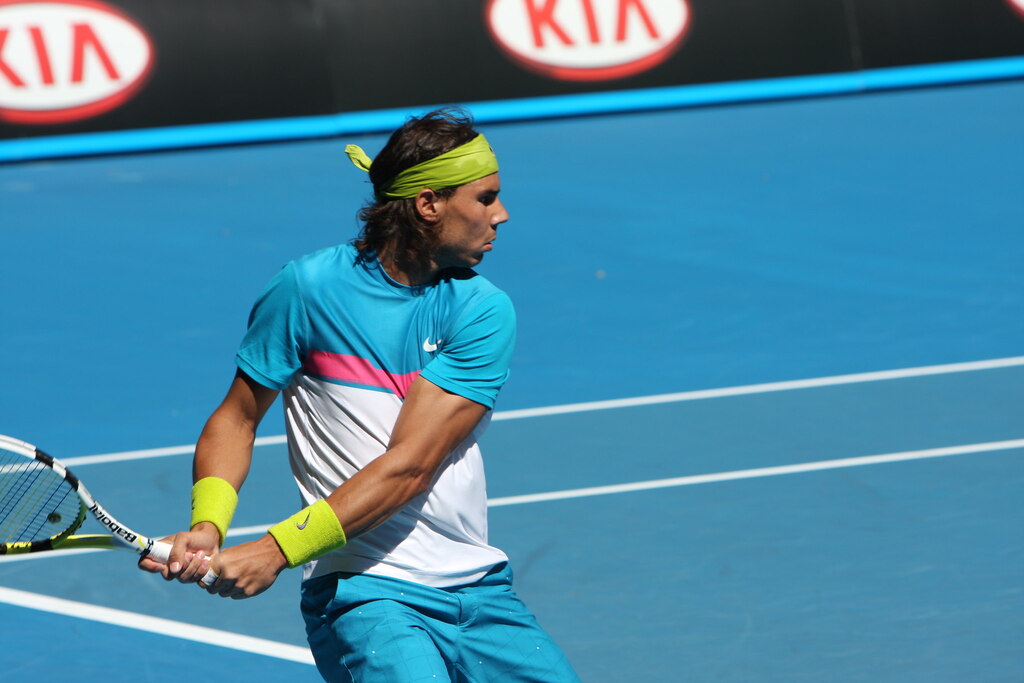 Jogador de tênis Rafael Nadal, usando roupa azul e branca, rebatendo uma bola durante a partida, em quadra azul.