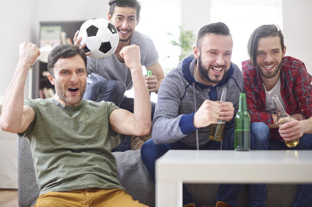 4 homens tomando cerveja e comemorando em um sofá, com um deles segurando uma bola de futebol.