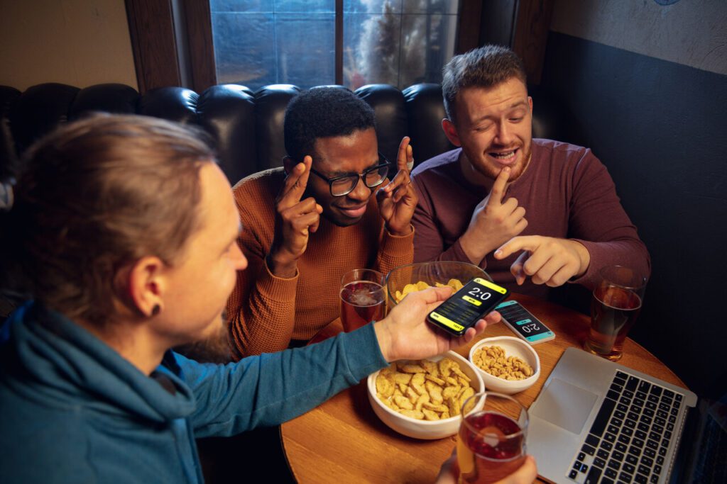 Três amigos sentados em volta de uma mesa redonda assistindo a um jogo no notebook. Eles estão ansiosos com o resultado.