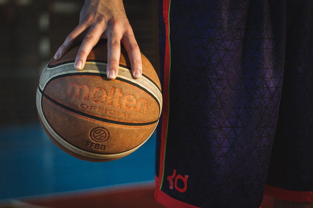 Jogador de basquete usando calção azul com detalhes vermelhos, segurado uma bola de basquete laranja com detalhes brancos,