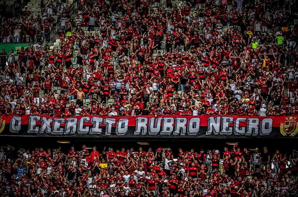 Torcida do Flamengo lotando arquibancada do estádio, usando uniforme do time e fazendo festa durante a partida.
