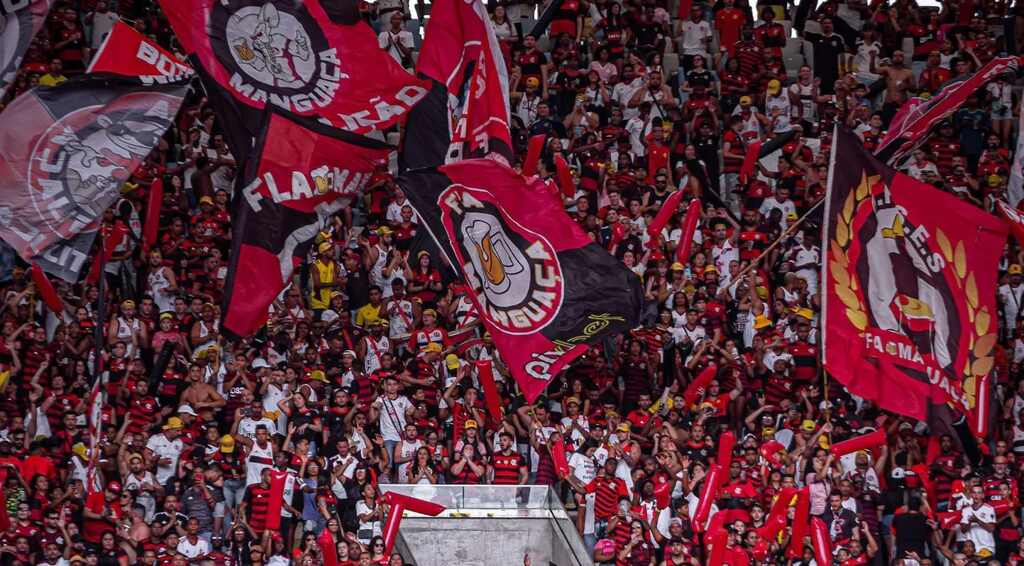 Torcida do Flamengo, vestindo uniforme preto e vermelho, levantando bandeiras e fazendo festa em estádio lotado.