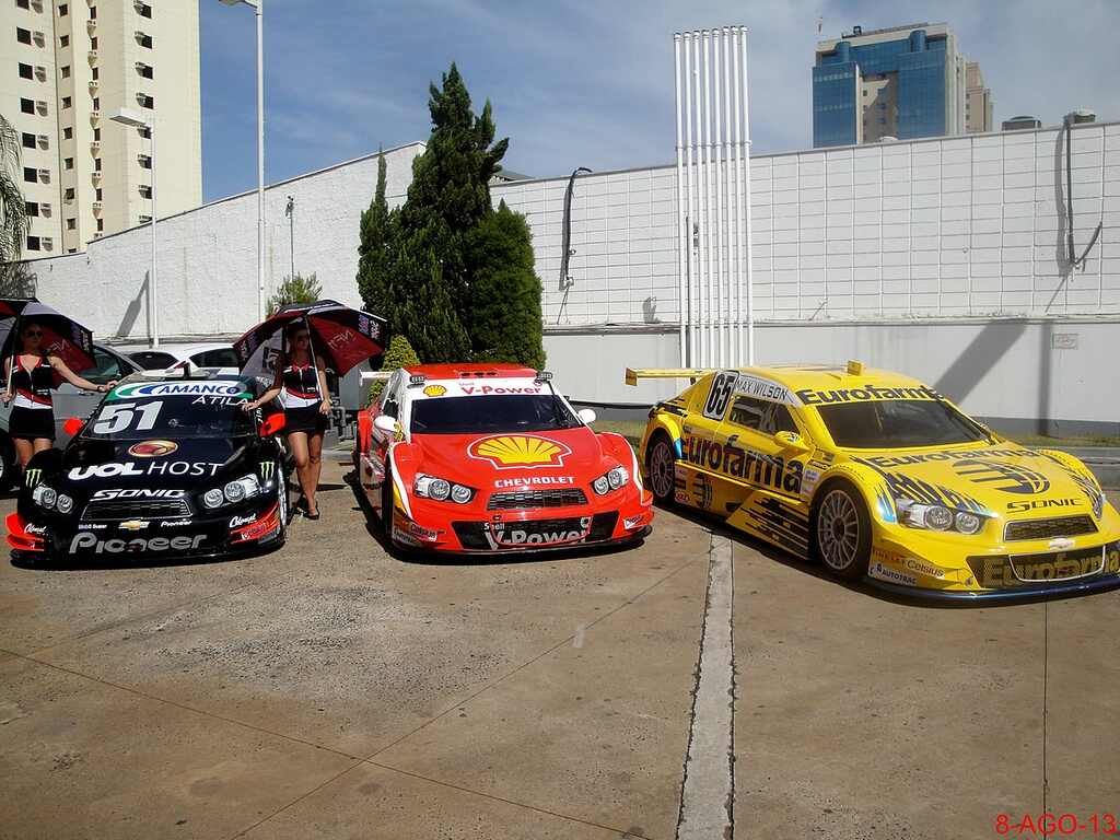 Tecnologia e inovações presentes nos carros da Stock Car 3 carros da Stock Car, um preto, um vermelho e um amarelo, deixados um ao lado durante, com duas mulheres tomando conta.