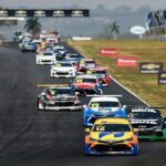 Diversos carros da Stock Car, de diferentes cores, enfileirados em pista de corrida durante trajeto.