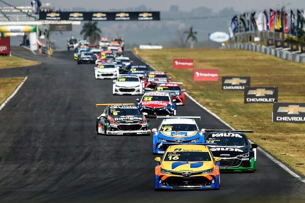 Diversos carros da Stock Car, de diferentes cores, enfileirados em pista de corrida durante trajeto.