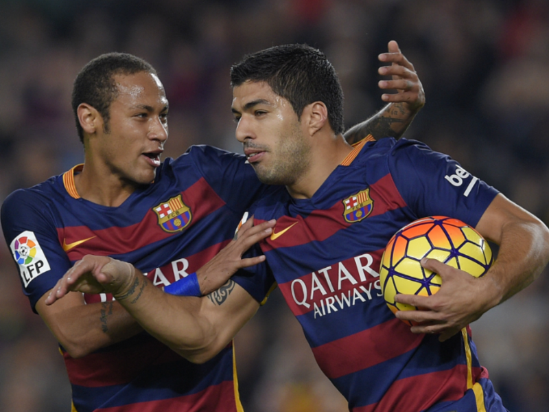 Luis Suárez e Neymar comemorando gol, campo de futebol com torcida vibrante ao fundo.