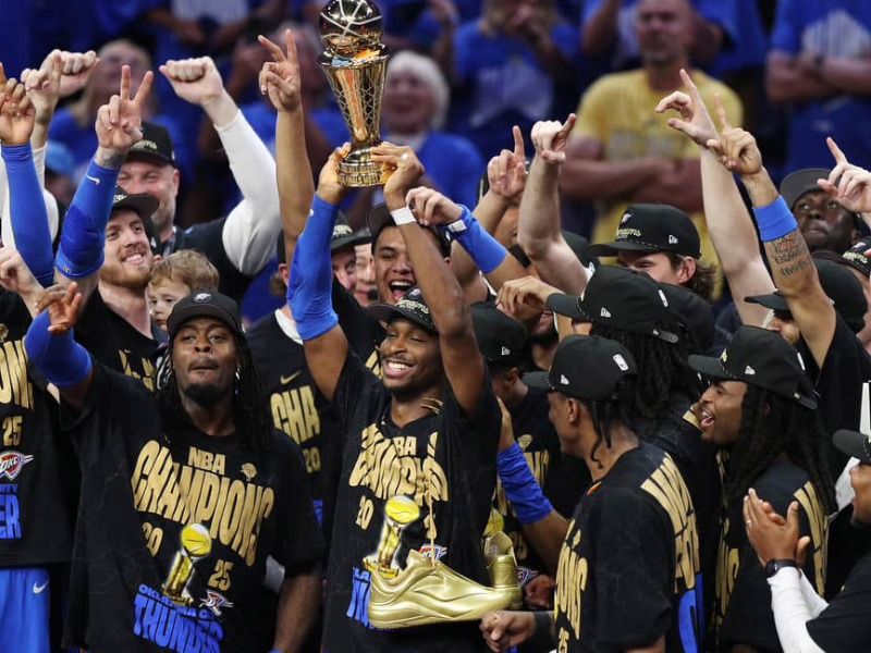 Shai Gilgeous-Alexander sorrindo com o troféu de MVP e a taça de campeão da NBA.