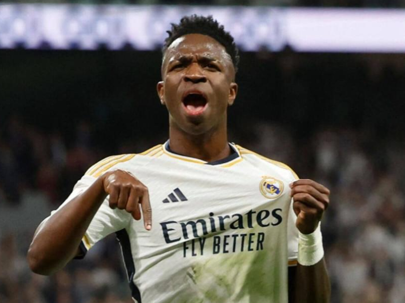 Close-up ultrarrealista de Vinícius Júnior, camisa 7 do Real Madrid, suado e sorridente, comemorando um gol em campo gramado sob a luz solar, transmitindo vitória e alívio.