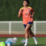 Zagueira de futebol feminino com o uniforme da seleção brasileira em campo, transmitindo alívio e determinação. Luz do sol realça o suor no rosto e a textura do gramado.