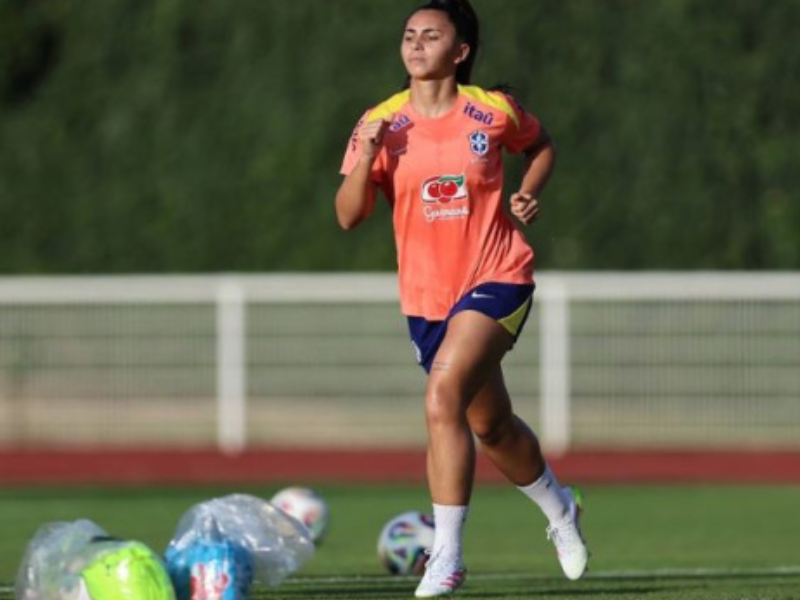 Zagueira de futebol feminino com o uniforme da seleção brasileira em campo, transmitindo alívio e determinação. Luz do sol realça o suor no rosto e a textura do gramado.