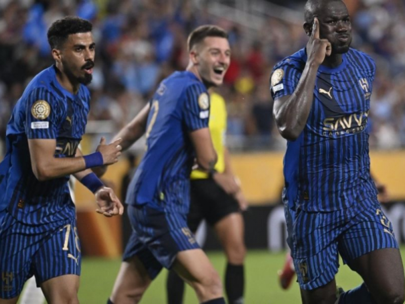 Jogador do Al Hilal em uniforme azul celebrando vitoriosamente em campo, com jogadores do Manchester City desapontados ao fundo.