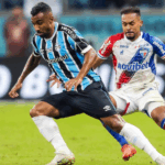 Campo de futebol durante um jogo noturno entre Grêmio e Fortaleza. Jogadores em ação, torcida ao fundo e iluminação artificial intensa.