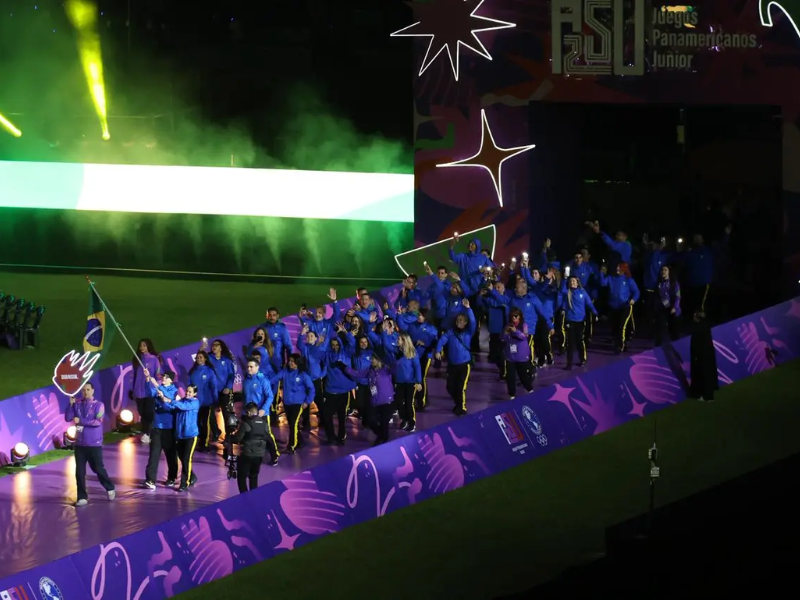 Cena ultrarrealista da cerimônia de abertura dos Jogos Pan-Americanos Júnior 2025 em um estádio, com atletas e bandeiras.