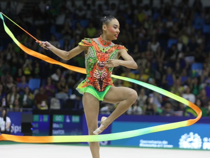 Duas ginastas rítmicas brasileiras, Bárbara Domingos e Geovanna Santos, em seus collants brilhantes, no palco de competição com torcedores ao fundo.