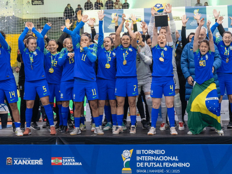 Jogadoras da Seleção Brasileira de Futsal Feminino comemorando um gol crucial contra a Colômbia, em quadra iluminada, com multidão ao fundo.