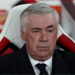 Retrato ultrarrealista de Carlo Ancelotti, técnico de futebol, em um campo de gramado sintético, vestindo terno azul, com o sol do fim da tarde e um estádio ao fundo. Sua expressão é de foco e sabedoria.